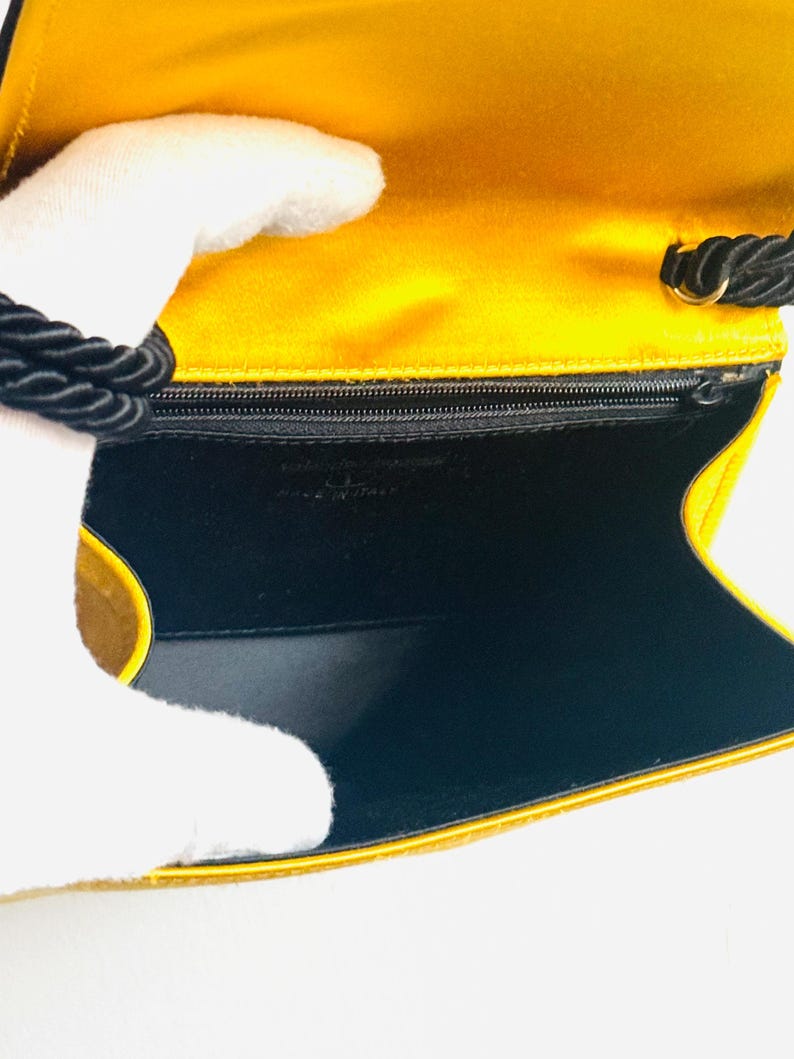 May include: Open view of a yellow satin clutch with a black rope handle. The interior is black with a small zipper pocket. The words "MADE IN ITALY" are visible.