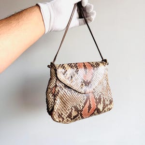 K&ouml;nnte beinhalten: Kleine Umh&auml;ngetasche mit Schlangenmuster in Braun-, Beige- und Korallt&ouml;nen. Die Tasche hat einen Klappverschluss und einen d&uuml;nnen, braunen Riemen. Die Tasche wird von einer behandschuhten Hand gehalten.