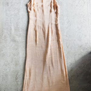 May include: A sleeveless, floor-length peach-colored dress. The dress has a scoop neckline and a fitted bodice that flows into a slightly flared skirt. The fabric has a subtle, textured pattern.