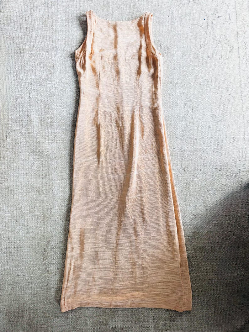 May include: A sleeveless, floor-length peach-colored dress. The dress has a scoop neckline and a fitted bodice that flows into a slightly flared skirt. The fabric has a subtle, textured pattern.
