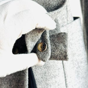 May include: Close-up of a gray wool coat. The coat features a buttoned cuff detail with a single brown button. The fabric has a textured appearance. The coat is being held by a gloved hand.