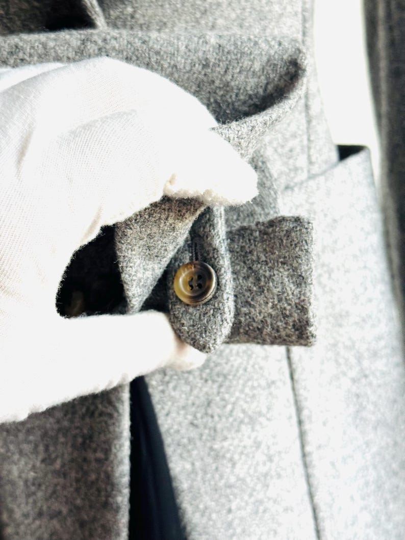 May include: Close-up of a gray wool coat. The coat features a buttoned cuff detail with a single brown button. The fabric has a textured appearance. The coat is being held by a gloved hand.