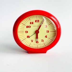 May include: Red alarm clock with a white face and black hands. The clock has black numbers and the text "WEHRL" and "OVET" printed on the face. The clock also has the text "MADE IN GERMANY" printed on the face.