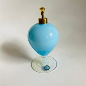 May include: A light blue glass perfume bottle with a gold metal top and a clear glass base. The bottle has a round body and a small, round base. The bottle is sitting on a white surface. The bottle has a sticker on the base that says "Made in Italy".