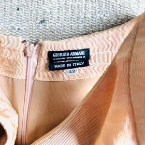 May include: Close-up of a peach-colored dress with a zipper and a label that reads "GIORGIO ARMANI MILANO - SUCCURSALE 29 MADE IN ITALY 42". The dress has a ribbed texture.