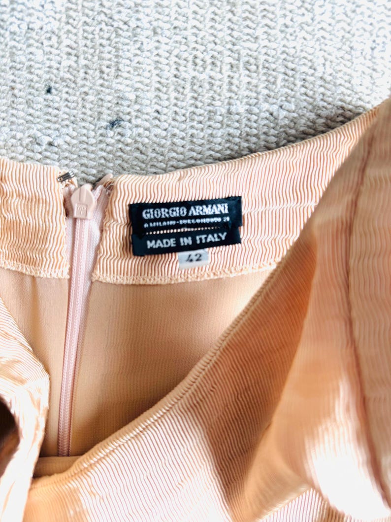 May include: Close-up of a peach-colored dress with a zipper and a label that reads "GIORGIO ARMANI MILANO - SUCCURSALE 29 MADE IN ITALY 42". The dress has a ribbed texture.