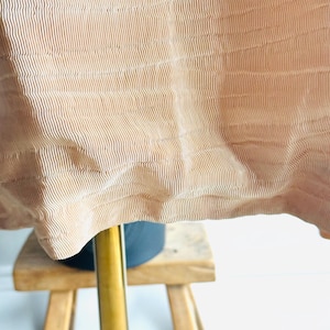 May include: Close-up of a tan fabric with vertical ribbed texture. The fabric drapes over a dark object and a wooden stool with three legs. The fabric has a wavy, flowing appearance.