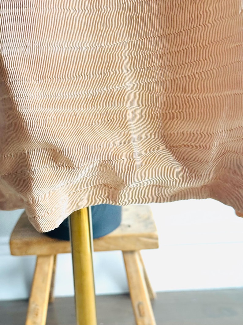 May include: Close-up of a tan fabric with vertical ribbed texture. The fabric drapes over a dark object and a wooden stool with three legs. The fabric has a wavy, flowing appearance.