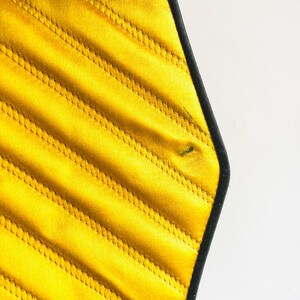 May include: Close-up of a yellow quilted bag with black trim. The bag has a geometric shape with diagonal stitching. The bag is made of a shiny material and has a black rope handle.