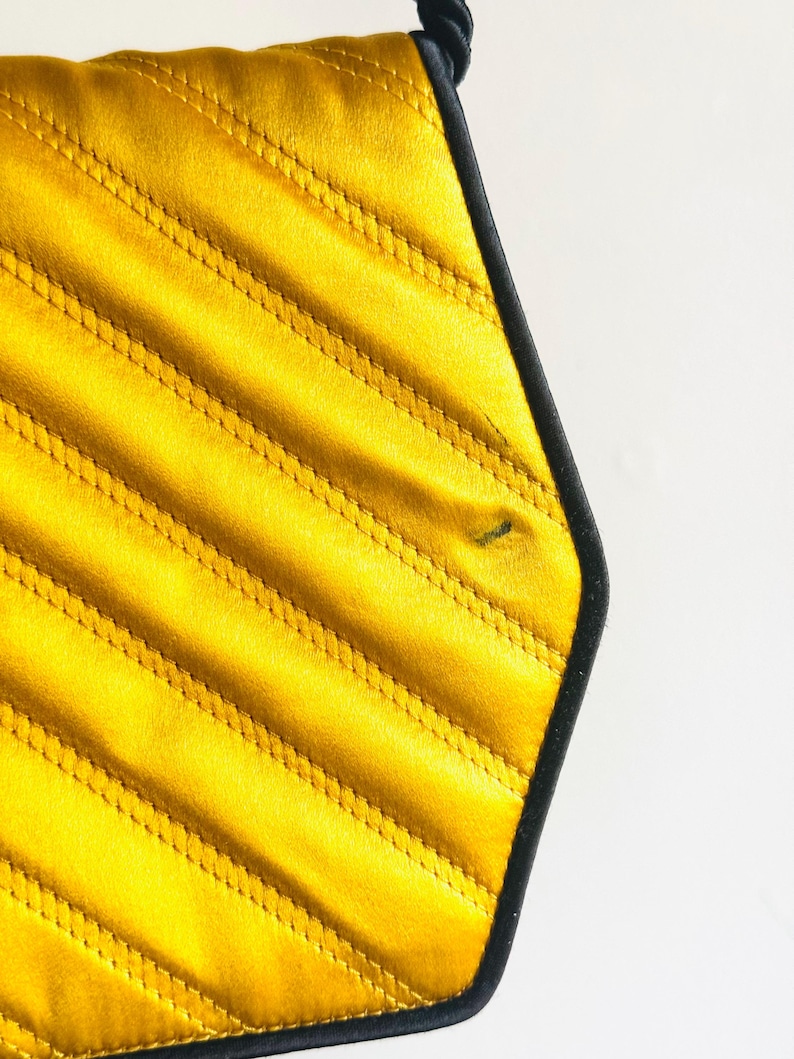 May include: Close-up of a yellow quilted bag with black trim. The bag has a geometric shape with diagonal stitching. The bag is made of a shiny material and has a black rope handle.