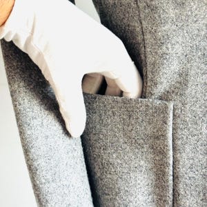 May include: Close-up of a gray wool coat with a pocket. A white-gloved hand is reaching into the pocket. The coat has a classic design and a textured surface. The coat is made of a soft, warm material.