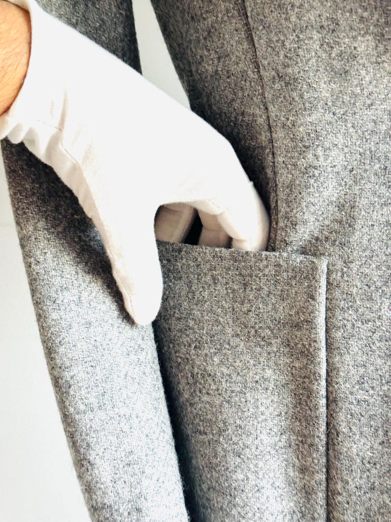 May include: Close-up of a gray wool coat with a pocket. A white-gloved hand is reaching into the pocket. The coat has a classic design and a textured surface. The coat is made of a soft, warm material.