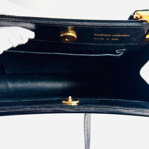 Może przedstawiać: Czarna sk&oacute;rzana torebka ze złotymi elementami. Wnętrze posiada małą kieszeń i napis "boutique valentino made in italy". Torba ma g&oacute;rny uchwyt i pasek na ramię.