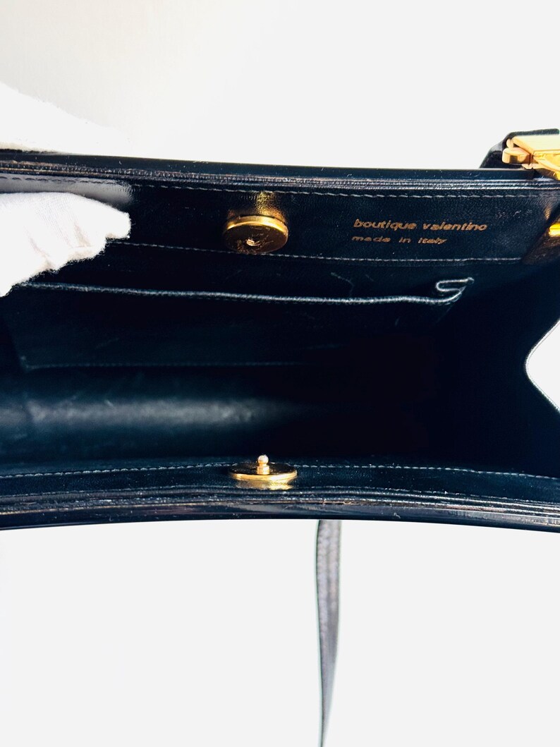 Może przedstawiać: Czarna sk&oacute;rzana torebka ze złotymi elementami. Wnętrze posiada małą kieszeń i napis "boutique valentino made in italy". Torba ma g&oacute;rny uchwyt i pasek na ramię.