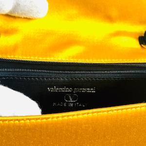 May include: Close-up of a yellow handbag interior. The lining is black with the words "valentino garavani" and "MADE IN ITALY" in silver lettering. The bag's exterior is a bright, shiny yellow.