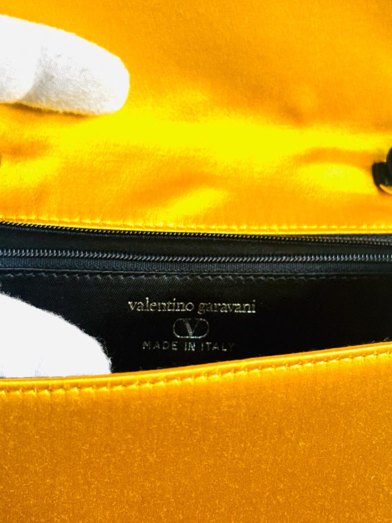 May include: Close-up of a yellow handbag interior. The lining is black with the words "valentino garavani" and "MADE IN ITALY" in silver lettering. The bag's exterior is a bright, shiny yellow.