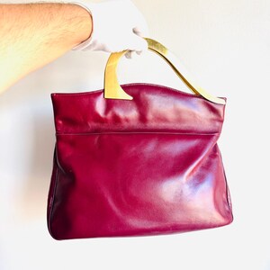 May include: A burgundy leather handbag with a gold-toned handle. The bag has a rectangular shape with a smooth, glossy finish. The handle is curved and attached to the top of the bag. The bag is held by a gloved hand.