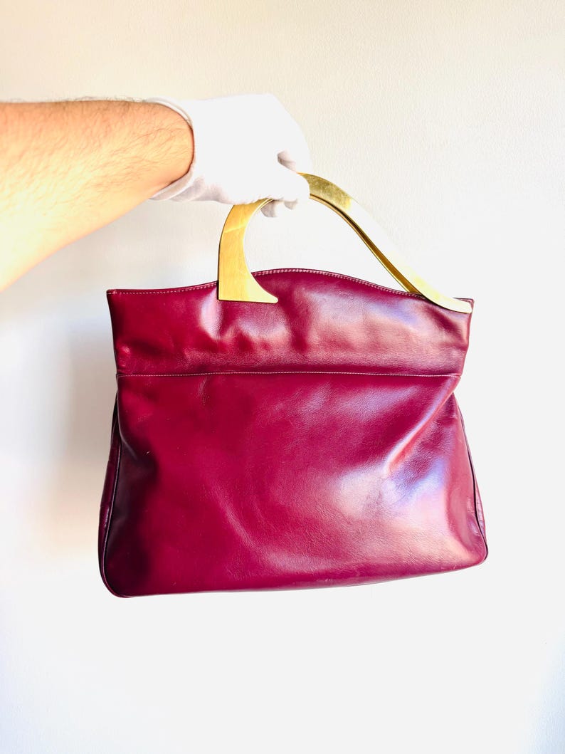 May include: A burgundy leather handbag with a gold-toned handle. The bag has a rectangular shape with a smooth, glossy finish. The handle is curved and attached to the top of the bag. The bag is held by a gloved hand.