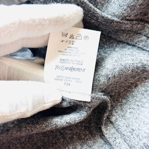 May include: Close-up of a gray wool garment with a tag. The tag displays care instructions and the Yves Saint Laurent logo, with "MADE IN ITALY" printed below. The garment's texture is visible.