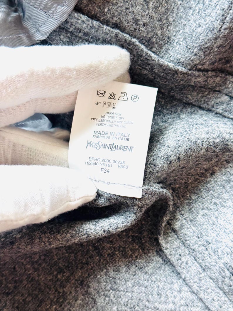 May include: Close-up of a gray wool garment with a tag. The tag displays care instructions and the Yves Saint Laurent logo, with "MADE IN ITALY" printed below. The garment's texture is visible.