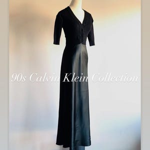 May include: A black vintage dress from the 90s Calvin Klein Collection. The dress features a short-sleeved, button-up top and a long, flowing skirt. The dress is displayed on a mannequin against a neutral background.