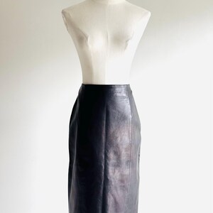 May include: A black leather pencil skirt displayed on a mannequin. The skirt is knee-length and has a classic, tailored design. The leather appears smooth and has a slight sheen. The mannequin is white and has a simple, minimalist design.