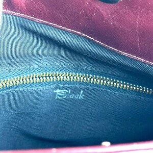May include: Burgundy leather handbag interior with a gold zipper and the word "Block" printed in white. The interior is lined with a dark blue fabric. The bag has a magnetic clasp.