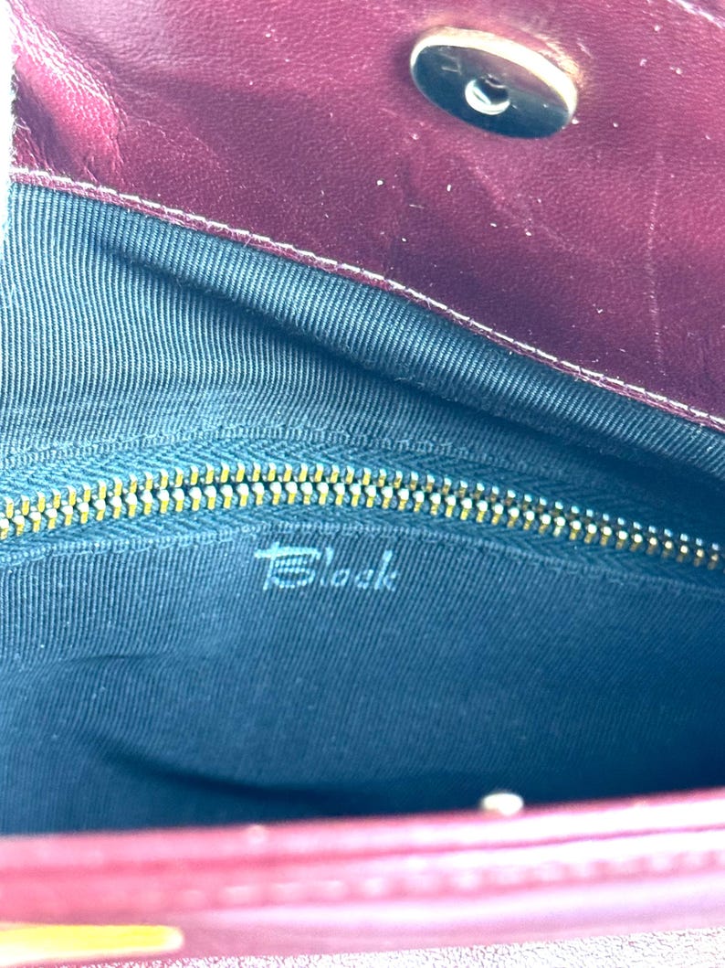 May include: Burgundy leather handbag interior with a gold zipper and the word "Block" printed in white. The interior is lined with a dark blue fabric. The bag has a magnetic clasp.