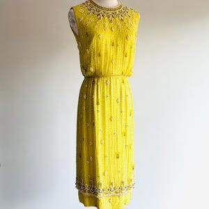 May include: A sleeveless, yellow dress with intricate beadwork. The dress features a beaded neckline, a cinched waist, and a knee-length skirt. The dress is adorned with white and silver beads, creating a shimmering effect. The dress is displayed on a mannequin.