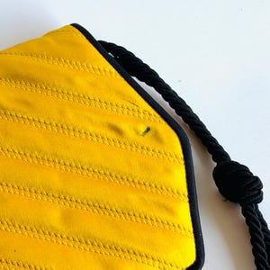 May include: A yellow quilted handbag with black trim and a black rope handle. The bag has a geometric shape and a textured surface. The handle is knotted at the end.