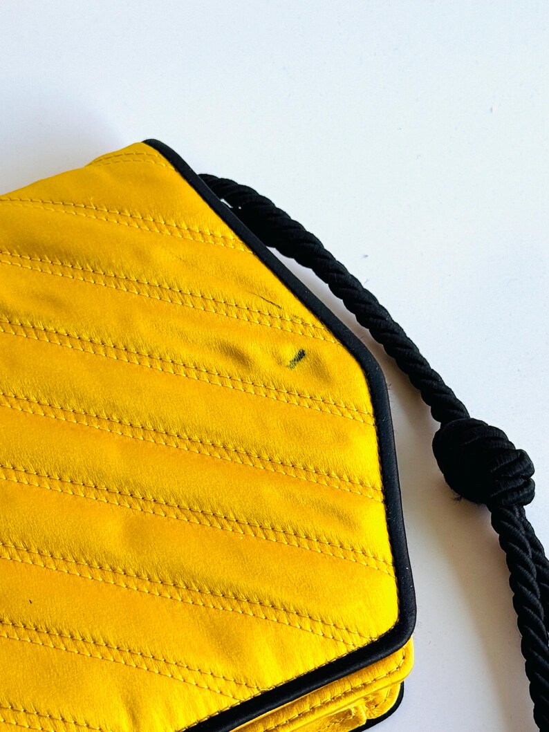 May include: A yellow quilted handbag with black trim and a black rope handle. The bag has a geometric shape and a textured surface. The handle is knotted at the end.