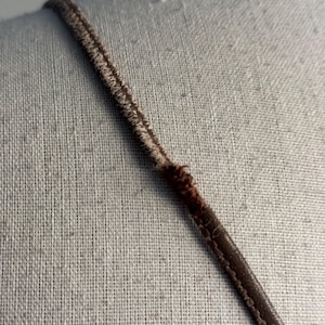 K&ouml;nnte beinhalten: Nahaufnahme eines braunen Riemens, m&ouml;glicherweise von einem Kleidungsst&uuml;ck, vor neutralem Hintergrund. Der Riemen besteht aus gen&auml;htem Material mit dunkelbraunen Kanten und einer helleren, strukturierten Mitte.