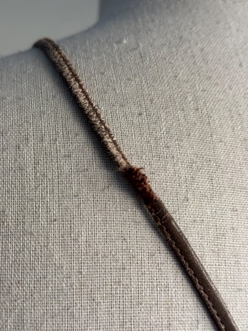 K&ouml;nnte beinhalten: Nahaufnahme eines braunen Riemens, m&ouml;glicherweise von einem Kleidungsst&uuml;ck, vor neutralem Hintergrund. Der Riemen besteht aus gen&auml;htem Material mit dunkelbraunen Kanten und einer helleren, strukturierten Mitte.