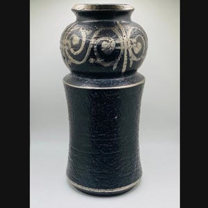 May include: A black ceramic vase with a textured, granular surface. The upper section features a rounded, bulbous shape adorned with silver-colored swirling patterns, while the lower body is cylindrical with a silver-colored rim at the base.