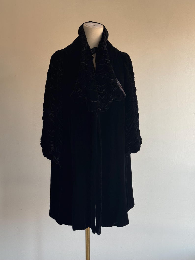 K&ouml;nnte beinhalten: Eine schwarze Samtjacke mit einem R&uuml;schenkragen und langen &Auml;rmeln. Die Jacke befindet sich auf einem Mannequin.