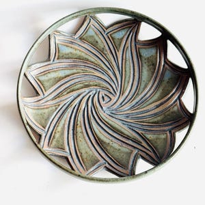 May include: A ceramic plate with a unique floral design. The plate features a spiral pattern with a light blue and green color scheme. The outer rim is a darker green, and the plate has a circular shape.