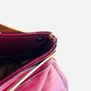 May include: A close-up of a burgundy leather handbag. The bag has a structured design with a flap closure. The interior is visible, revealing a dark lining. The bag has a gold-colored accent on the side.