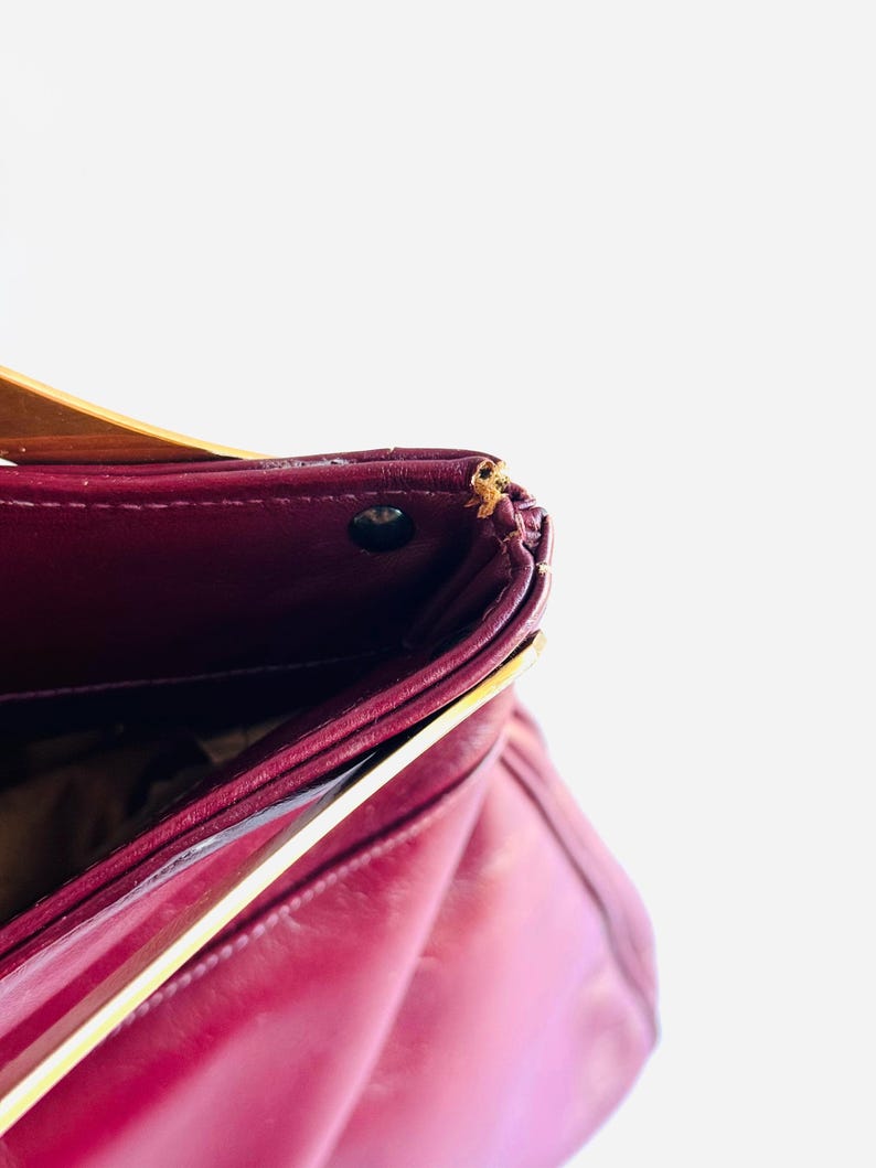 May include: A close-up of a burgundy leather handbag. The bag has a structured design with a flap closure. The interior is visible, revealing a dark lining. The bag has a gold-colored accent on the side.