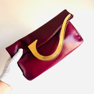 May include: A burgundy leather clutch with a gold-toned handle. The clutch has a curved, semi-circular handle and a rectangular shape. The leather has a glossy finish. The bag is held by a gloved hand.
