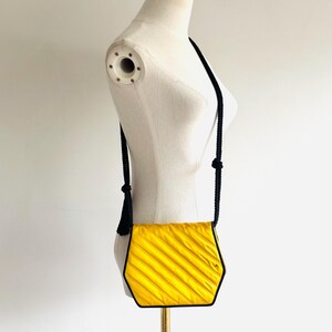 May include: A yellow, hexagonal crossbody bag with a black trim and strap. The bag has a textured, pleated design. The strap is made of black rope and has a tassel on one end. The bag is displayed on a mannequin.