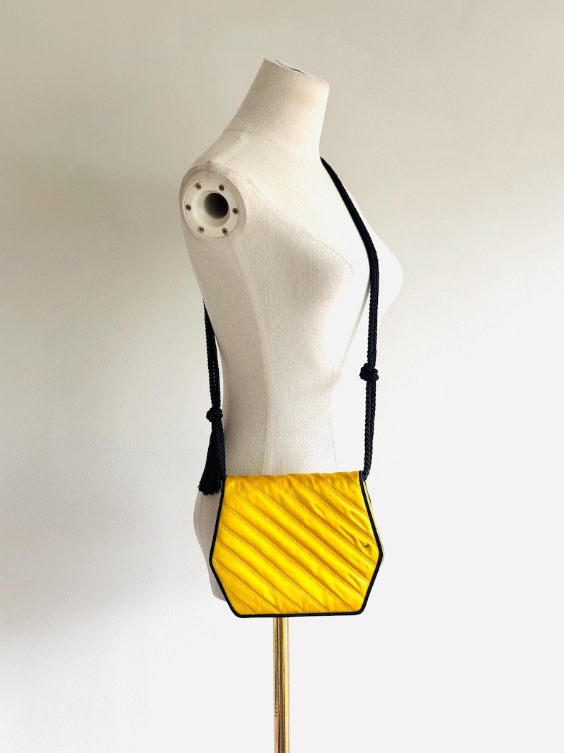 May include: A yellow, hexagonal crossbody bag with a black trim and strap. The bag has a textured, pleated design. The strap is made of black rope and has a tassel on one end. The bag is displayed on a mannequin.