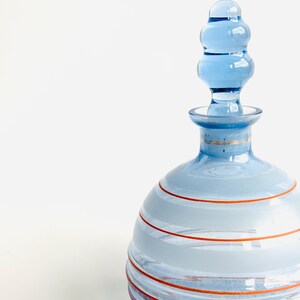 Pu&ograve; includere: Una bottiglia di vetro blu con un design a strisce e un tappo di vetro trasparente. La bottiglia ha un corpo rotondo e un collo sottile. Le strisce sono di colore blu chiaro e arancio.