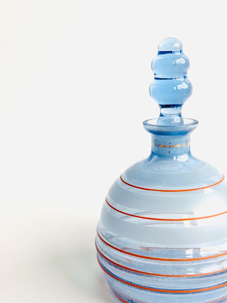 Pu&ograve; includere: Una bottiglia di vetro blu con un design a strisce e un tappo di vetro trasparente. La bottiglia ha un corpo rotondo e un collo sottile. Le strisce sono di colore blu chiaro e arancio.