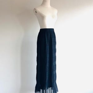 May include: A long, black skirt with a textured pattern and a ruffled hem. The skirt is displayed on a mannequin against a white background. The skirt has an elastic waistband.