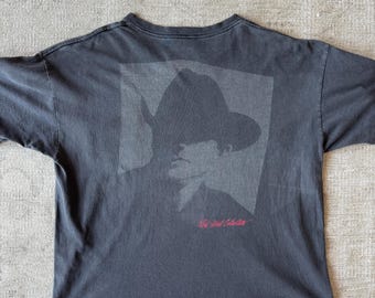 Op de afbeelding: Donkergrijs t-shirt met een vervaagde silhouet van een persoon met hoed en pak. De tekst "Wild West Theater" is in het rood onder de afbeelding gedrukt. Het shirt is gemaakt van een zacht, versleten materiaal.