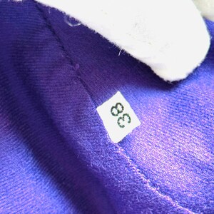 Pode incluir: Close-up de um tecido roxo vibrante com uma textura vis&iacute;vel. Uma pequena etiqueta branca com o n&uacute;mero "38" em preto est&aacute; presa ao tecido. O tecido &eacute; parcialmente obscurecido por um objeto branco.