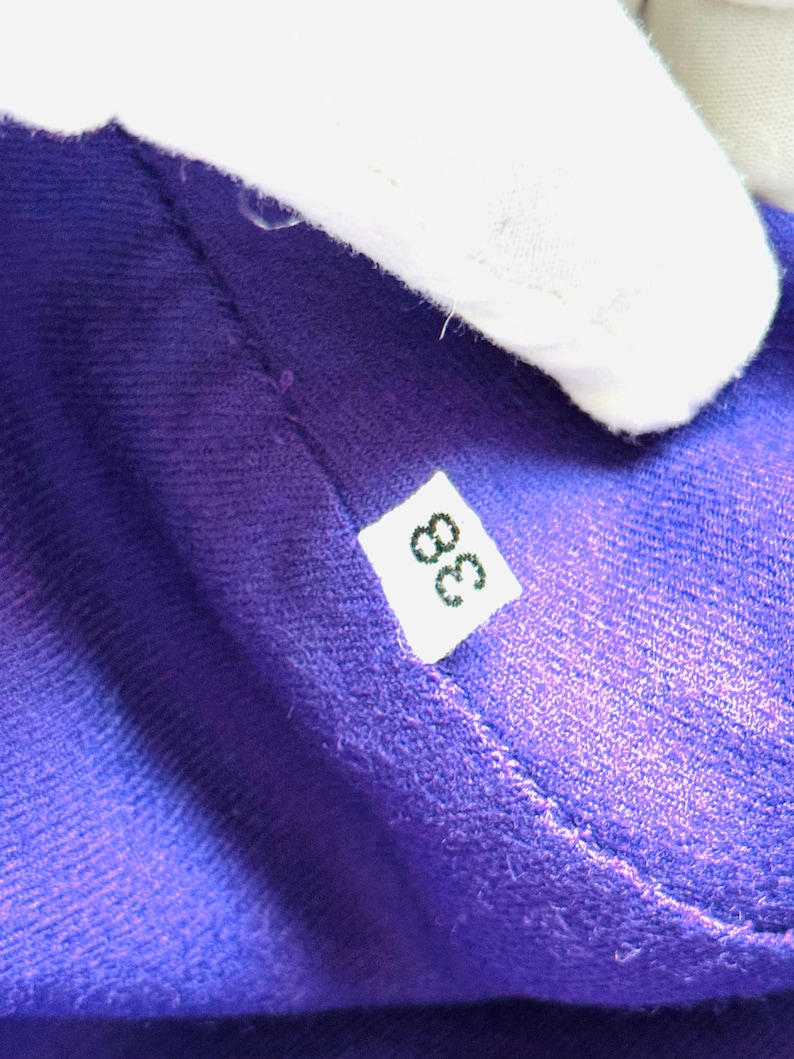 Pode incluir: Close-up de um tecido roxo vibrante com uma textura vis&iacute;vel. Uma pequena etiqueta branca com o n&uacute;mero "38" em preto est&aacute; presa ao tecido. O tecido &eacute; parcialmente obscurecido por um objeto branco.