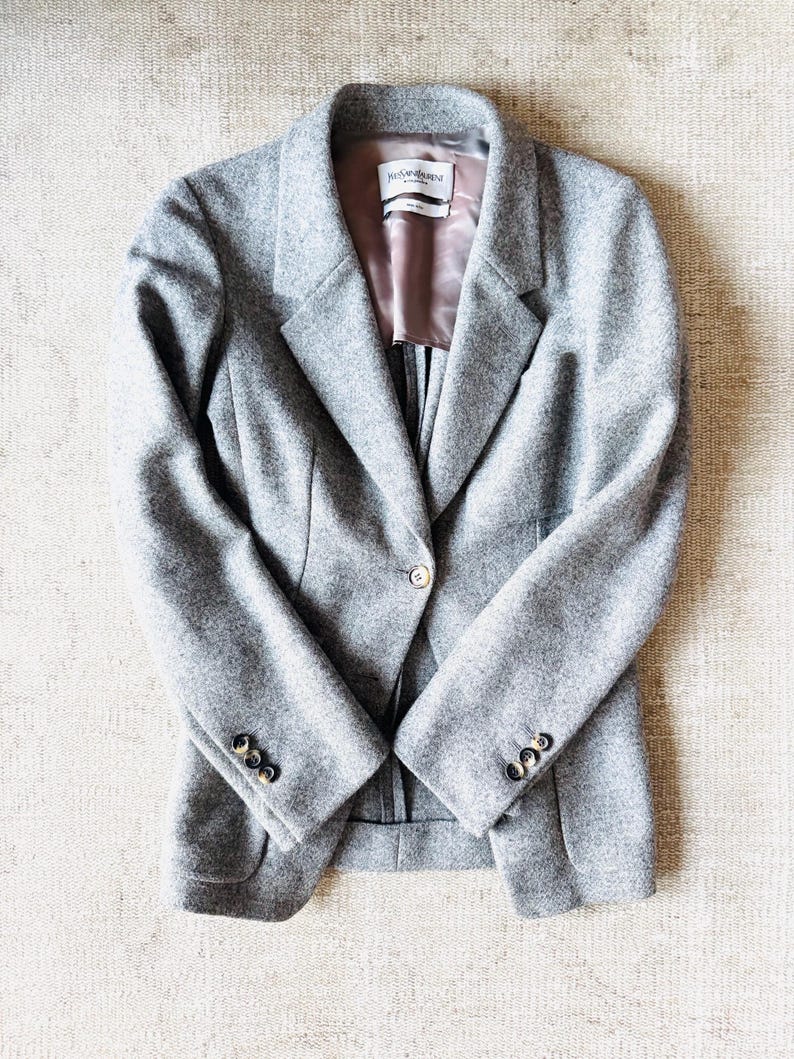 May include: A gray wool blazer with a single-button closure and three buttons on each sleeve. The blazer has a notched lapel and a satin lining. The label inside the blazer reads "Yves Saint Laurent".