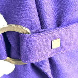 Pode incluir: Close-up de um tecido roxo vibrante com uma fivela met&aacute;lica e um pequeno detalhe quadrado dourado. A fivela tem texto gravado. O tecido &eacute; dobrado, criando uma apar&ecirc;ncia texturizada.
