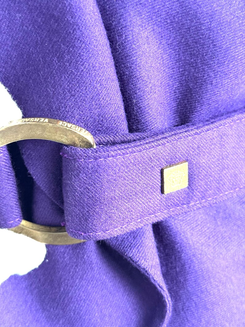 Pode incluir: Close-up de um tecido roxo vibrante com uma fivela met&aacute;lica e um pequeno detalhe quadrado dourado. A fivela tem texto gravado. O tecido &eacute; dobrado, criando uma apar&ecirc;ncia texturizada.